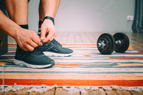 Close-up of a man tying shoelaces in black sports sneakers, a sports black watch on his hand, and metal dumbbells nearby © Виталий Сова