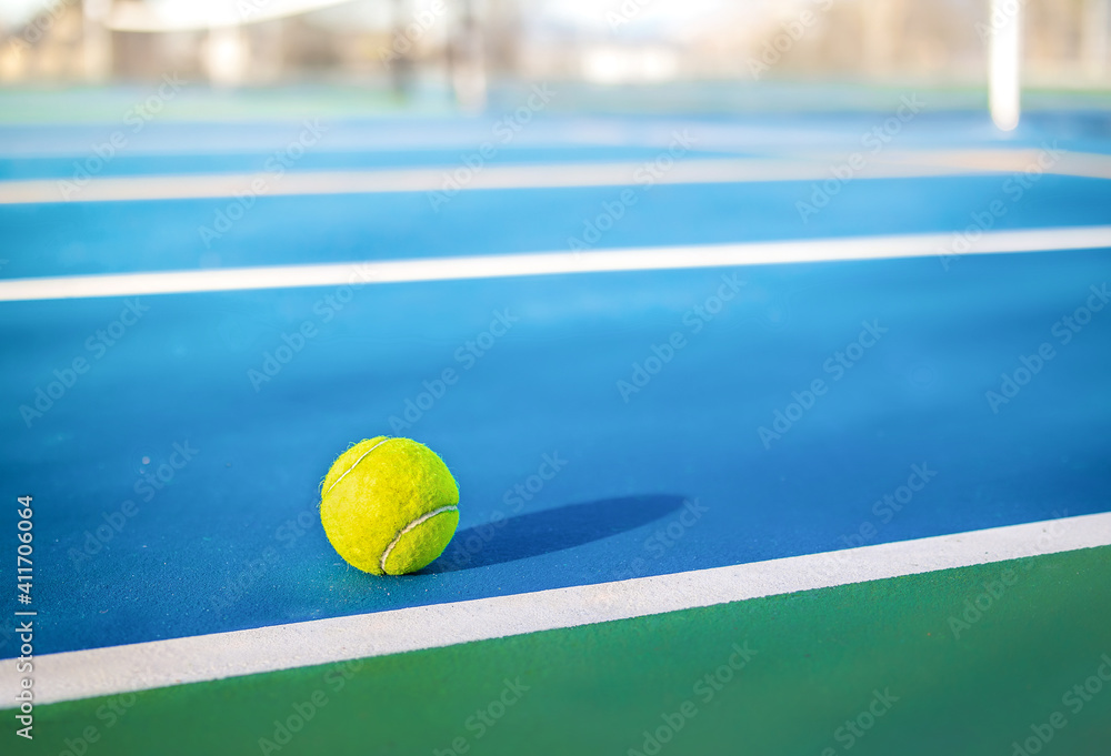 Yellow tennis ball next to sideline in outdoor tennis court, closeup. Defocused and abstract blue and green rubberized ground surface for shock absorption. Tennis sport texture. Selective focus.