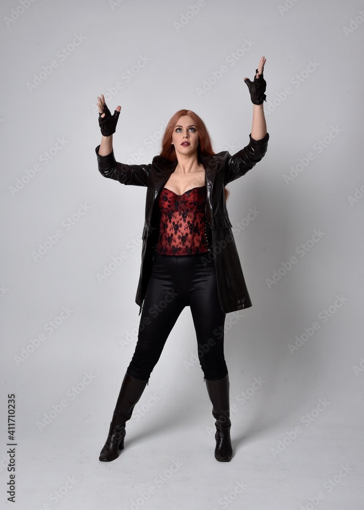 full length portrait of girl with long red hair wearing dark leather coat, corset and boots. Standing pose facing front on with magical hand gestures against a studio background. full length portrait of girl with long red hair wearing dark leather coat, corset and boots. Standing pose facing front on with magical hand gestures against a studio background.