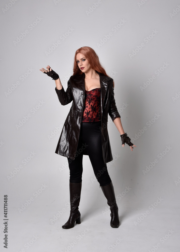 full length portrait of girl with long red hair wearing dark leather coat, corset and boots. Standing pose facing front on with magical hand gestures against a studio background. full length portrait of girl with long red hair wearing dark leather coat, corset and boots. Standing pose facing front on with magical hand gestures against a studio background.