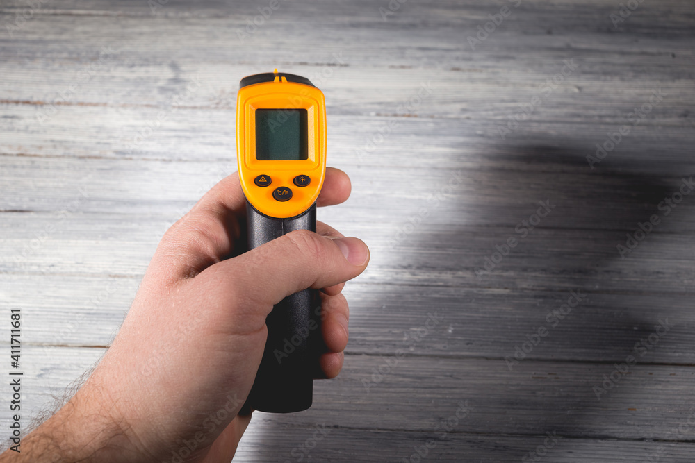 Fototapeta premium A man holds a yellow-black pyrometer on a wooden background. A device for non-contact temperature measurement.