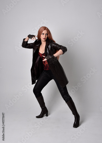full length portrait of girl with long red hair wearing dark leather coat, corset and boots. Standing pose facing front on with magical hand gestures against a studio background.