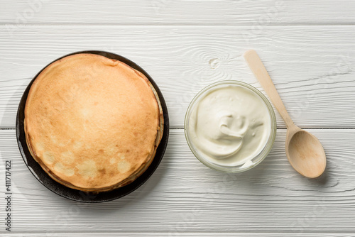 Flat lay composition with a plate of pancakes, sour cream and a wooden spoon on the table.