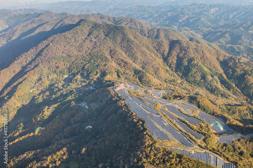 ソーラーパネルの傍を飛ぶパラグライダーと紅葉に染まる龍門山を空撮。和歌山県紀の川市 © NCP