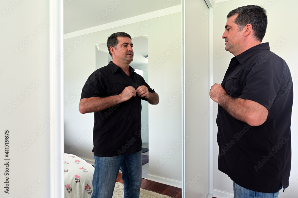 Middle aged man (age 40-50) looking at mirror buttoning up a shirt getting dressing up in home bedroom. Real people. Copy space Middle aged man (age 40-50) looking at mirror buttoning up a shirt getting dressing up in home bedroom. Real people. Copy space