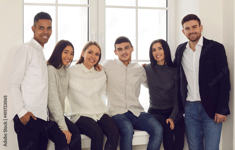 Group portrait of cheerful and stylish multiracial students sitting on the windowsill and hugging looking at the camera. Concept of education, friendship and unity. Group portrait of cheerful and stylish multiracial students sitting on the windowsill and hugging looking at the camera. Concept of education, friendship and unity.