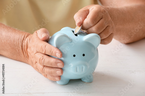 Senior woman putting coin into piggy bank at table. Concept of pension