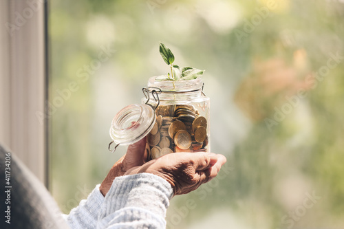 Senior woman with savings and young plant in jar near window at home. Concept of pension