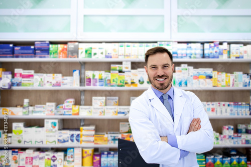 Portrait of handsome pharmacist standing in drugstore with shelf full of medicines in background.