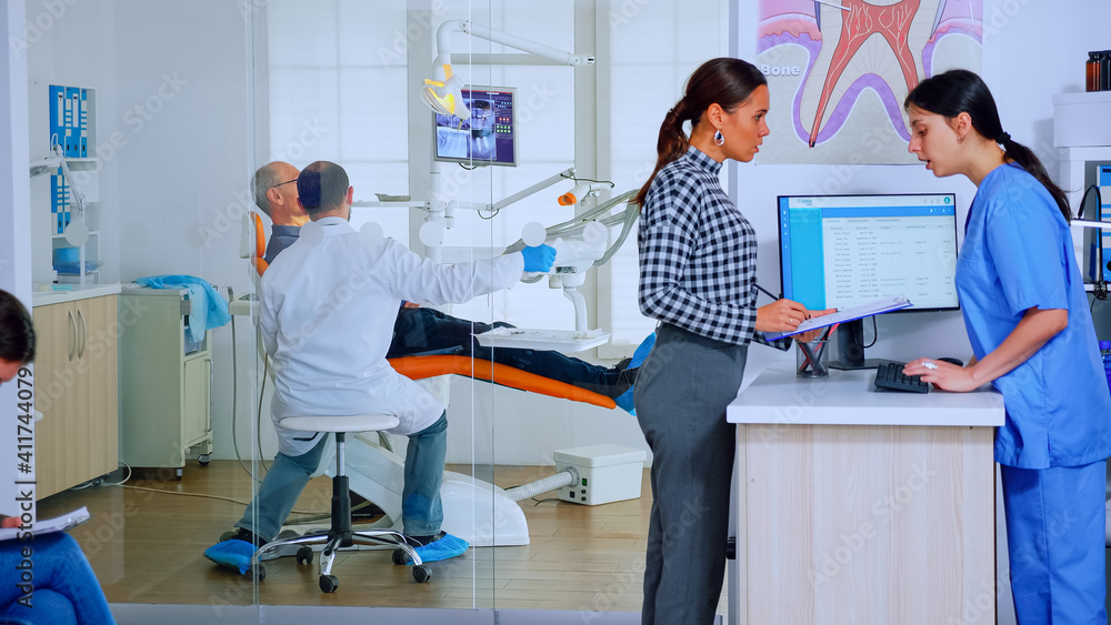 Obraz premium Patients asking informations filling in dental document preparing for teeth exemination. Senior woman sitting on chair in waiting area of crowded orthodontist office while doctor working in background