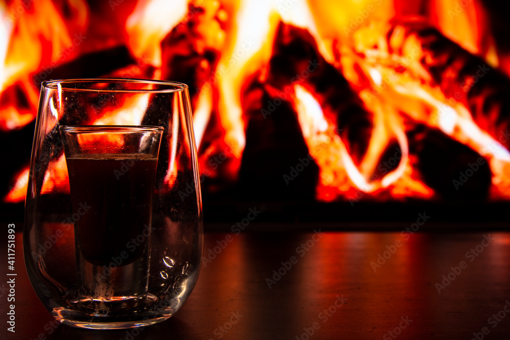Fototapeta premium A beautiful glass with a drink near the fireplace. Firewood and fire. Glass and liquid.