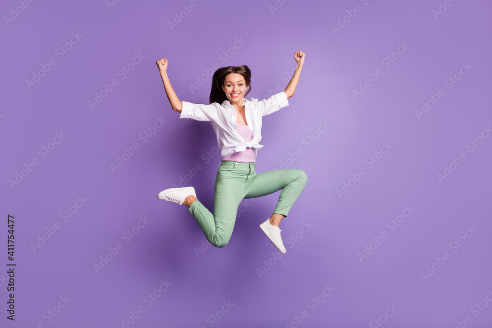 Full length photo of funny lady jump raise fists have fun wear pants footwear white shirt isolated violet color background Full length photo of funny lady jump raise fists have fun wear pants footwear white shirt isolated violet color background