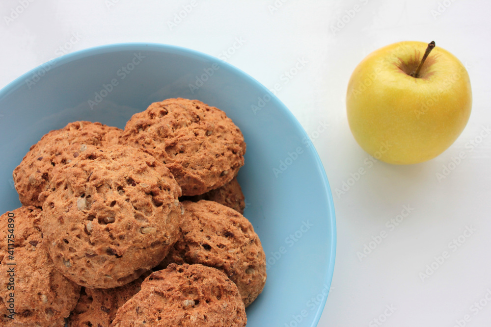 Homemade oatmeal cookies in a blue bowl and golden apple on white table. Overhead view of oat biscuits and fresh fruits on white background with copy space. Homemade oatmeal cookies in a blue bowl and golden apple on white table. Overhead view of oat biscuits and fresh fruits on white background with copy space.