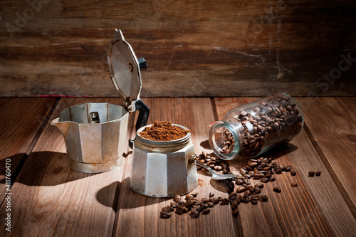 A geyser type metal coffee maker, photographed against a vintage wooden background with coffee beans.
