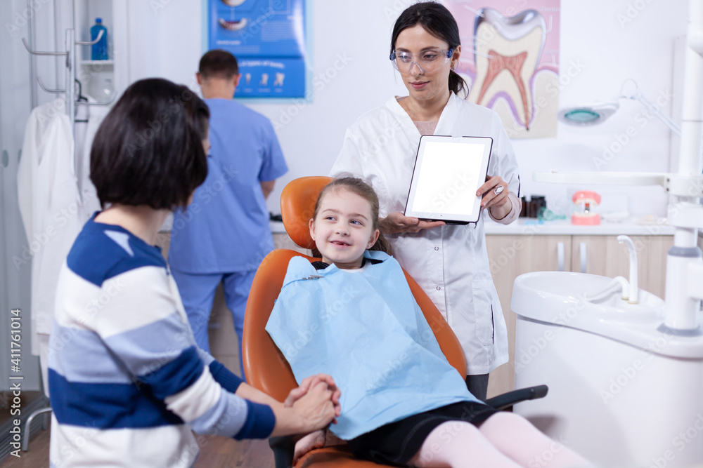 Obraz premium Little girl wearing dental bib in the course of dentistiry visit with parent and doctor holding device with white screen. Stomatolog explaining teeth prevention to mother and child holding tablet pc