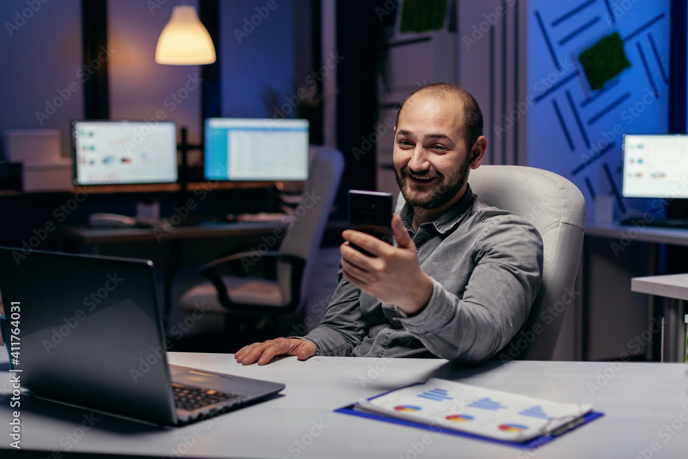 Happy freelancer sitting at desk discussing on online call using smartphone. Businessman in the course of an important video conference while doing overtime at the office. Happy freelancer sitting at desk discussing on online call using smartphone. Businessman in the course of an important video conference while doing overtime at the office.