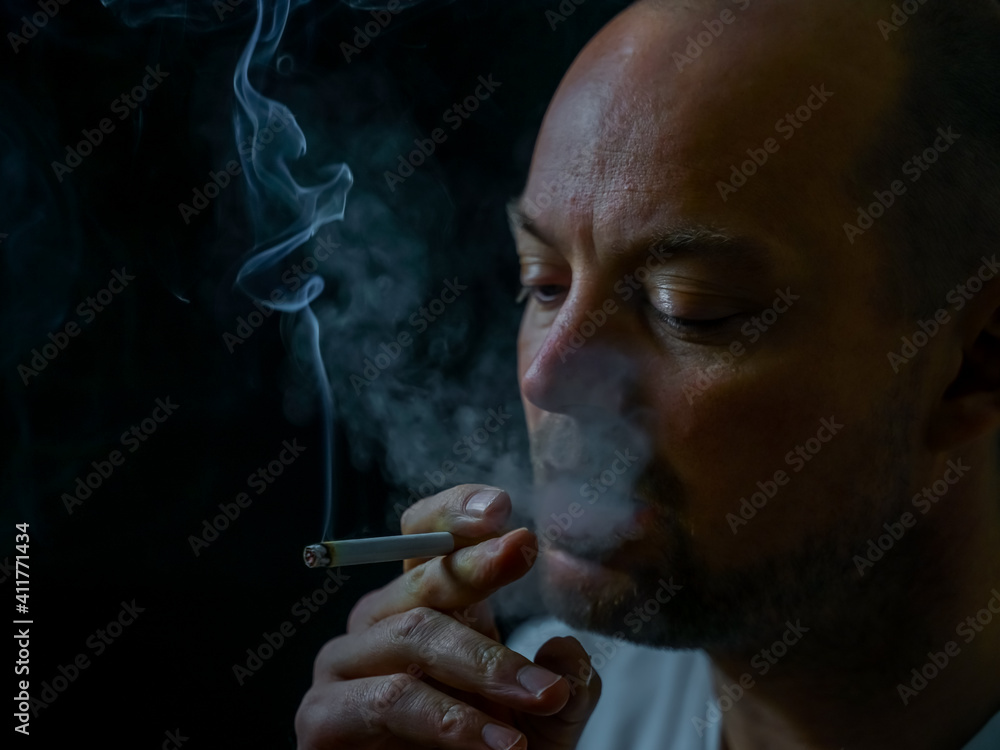 Fototapeta premium Man smoking a cigarette blowing out white smoke. Isolated on black background, shallow depth of field.