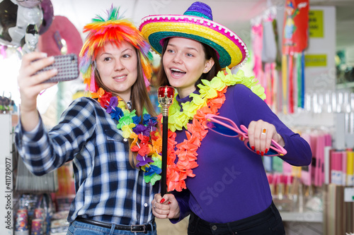Comically dressed happy girls making funny selfies photo in festive accessories shop
