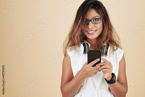 Portrait of cheerful young Asian woman in glasses choosing music on smartphone to listen to in headphones