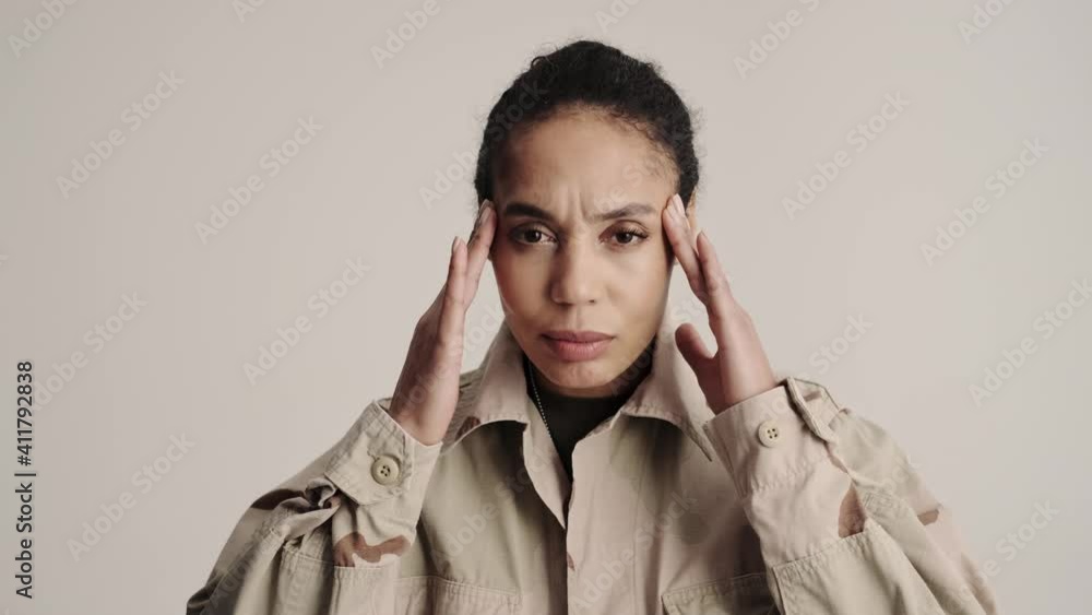A close-up view of a thinking young american soldier woman is massaging her temples standing isolated over a gray background in the studio A close-up view of a thinking young american soldier woman is massaging her temples standing isolated over a gray background in the studio