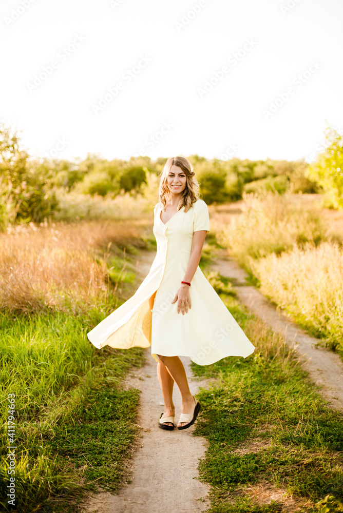 A young beautiful girl with blond curly hair in a light summer yellow dress and with a straw hat in her hands in a field in the countryside at sunset. People and nature A young beautiful girl with blond curly hair in a light summer yellow dress and with a straw hat in her hands in a field in the countryside at sunset. People and nature