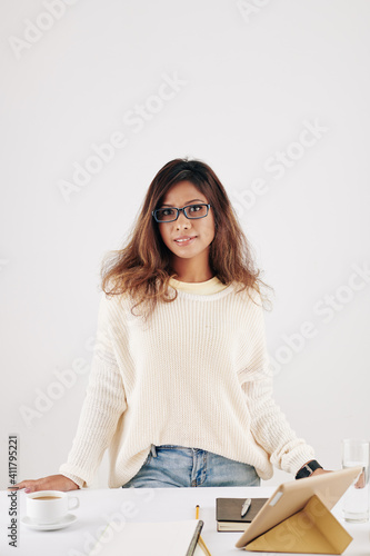 Portrait of smiling attractive young Asian woman in sweater standing at desk with cup of coffee, tablet computer and textbooks prepared for online class