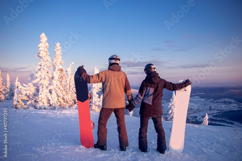 Loving couple of snowboarders hold hands and look into distance, rear view. Winter forest with snow, sunset