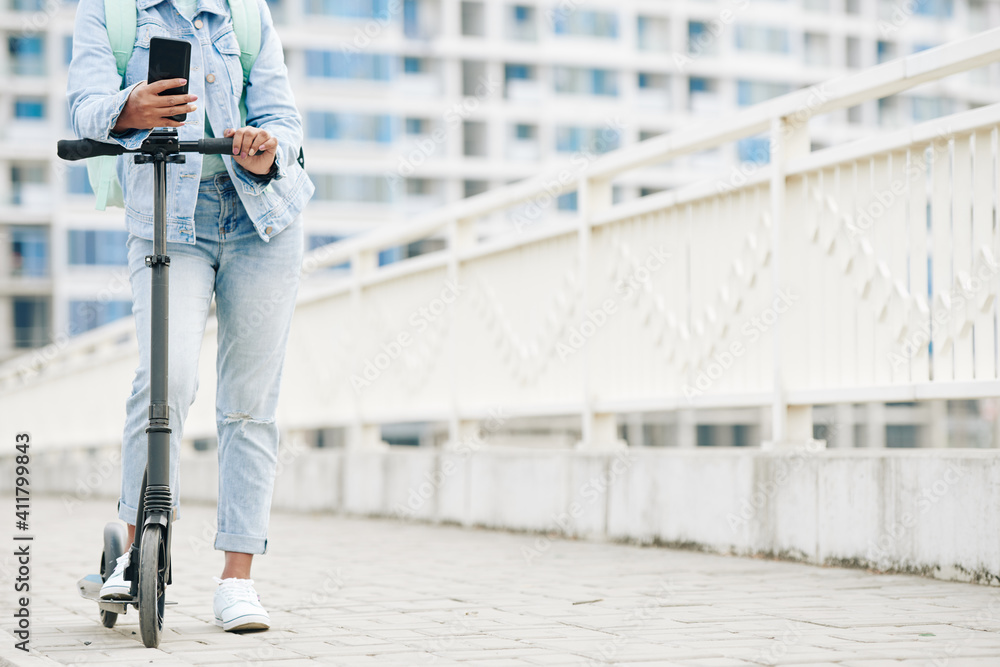 Cropped image of young woman in jeans standing on electric scooter with smartphone in hands