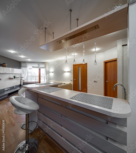 Contemporary interior of kitchen and living room in luxury flat. New kitchen set with light wooden counter. Wooden doors. Bar stool.