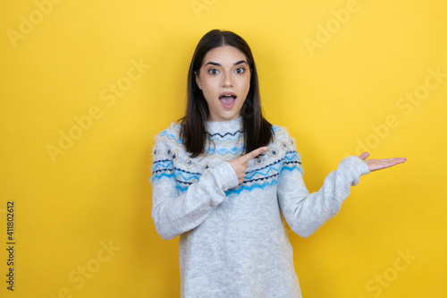 Young caucasian woman wearing casual sweater over yellow background surprised, showing and pointing something that is on her hand