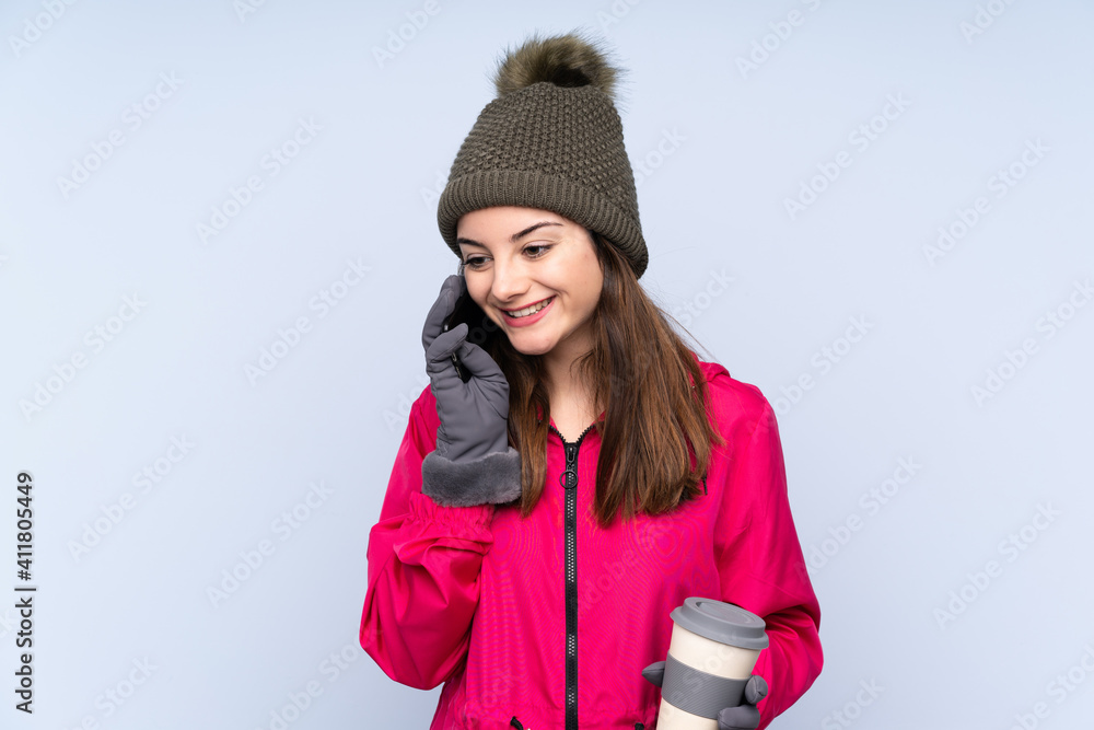 Young girl with winter hat isolated on blue background holding coffee to take away and a mobile Young girl with winter hat isolated on blue background holding coffee to take away and a mobile