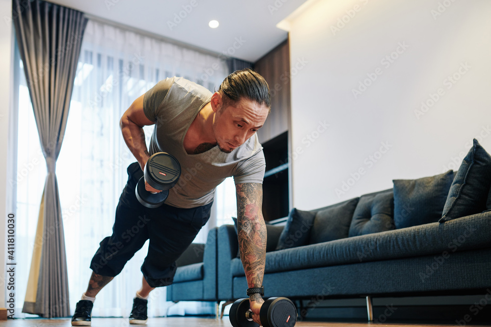 Serious fit mixed-race man doing dumbbells plank row exercise lifting dumbbell weights in his apartment