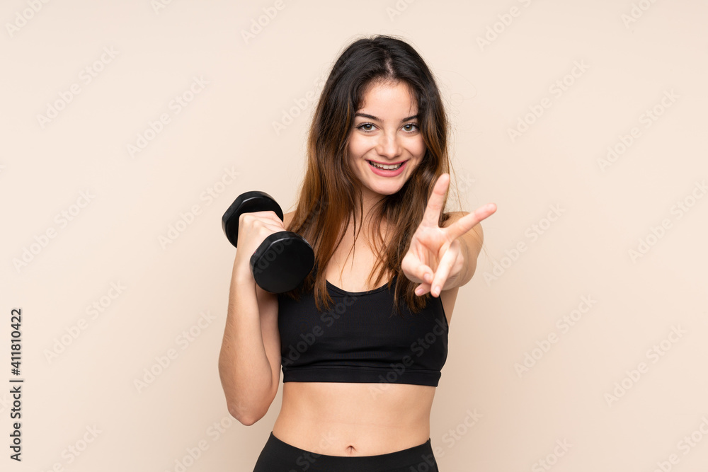 Young sport woman making weightlifting isolated on beige background smiling and showing victory sign Young sport woman making weightlifting isolated on beige background smiling and showing victory sign