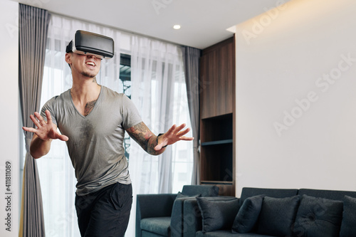 Excited young man in virtual reality headset playing video game at home, trying to entertain himself at home