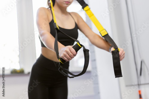 Young fit woman trains muscles with trx resistance belts in light room