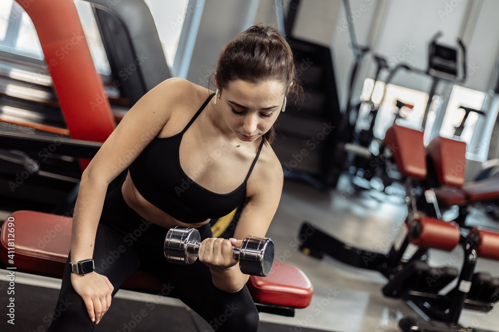 Fit woman training biceps, doing concentrated lifting dumbbell for biceps while sitting on a bench in the gym Fit woman training biceps, doing concentrated lifting dumbbell for biceps while sitting on a bench in the gym