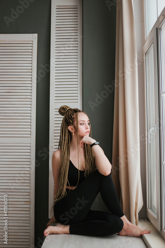 Young woman with long braids in sportswear practices yoga. A beautiful girl is sitting near a large window.