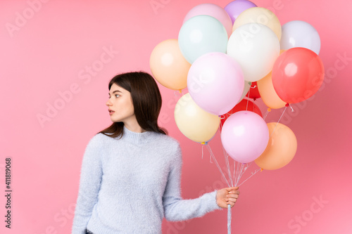 Young Ukrainian teenager girl holding lots of balloons over isolated pink background looking side