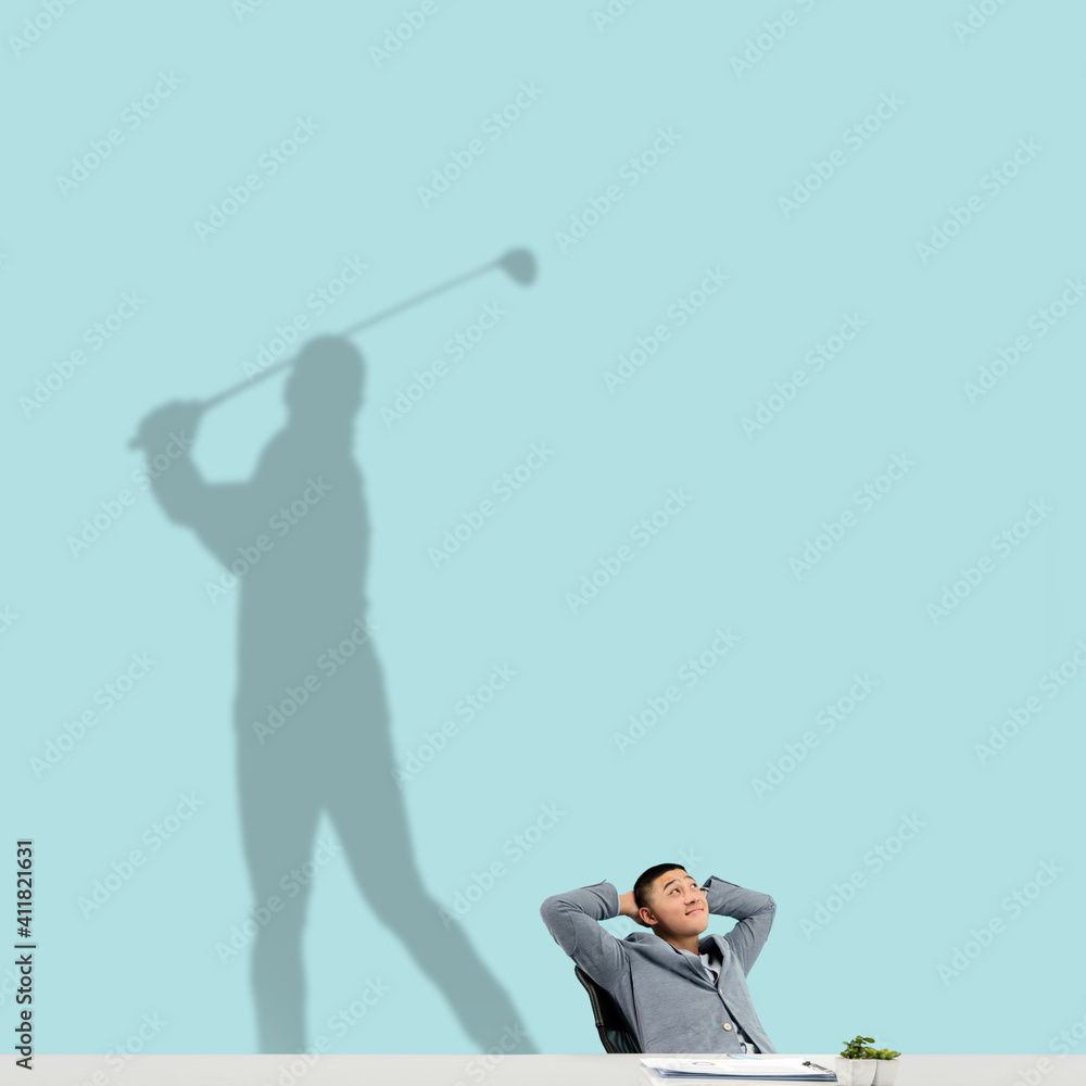 Young asian man dreaming about future in big sport during his work in office. Becoming a legend. Shadow, silhouette of professional golf player on the wall. Inspiration, aspiration. Copyspace.