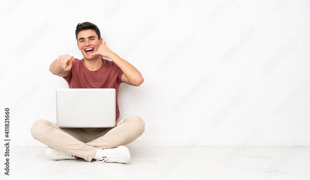 Teenager man sitting on the flor with his laptop making phone gesture and pointing front Teenager man sitting on the flor with his laptop making phone gesture and pointing front