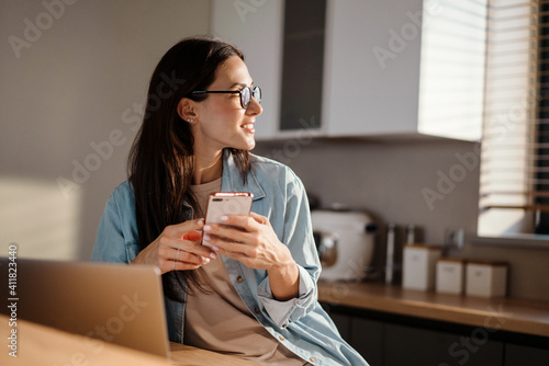 Happy charming woman using smartphone while working with laptop
