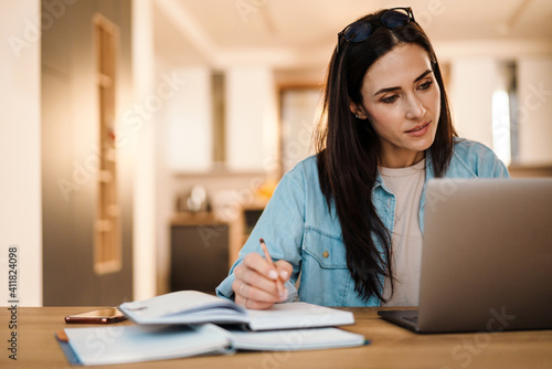 Focused charming woman writing down notes while working with laptop