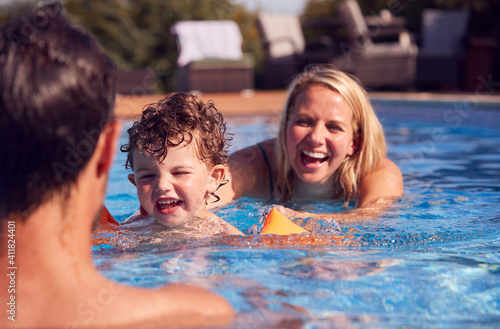 Family In Outdoor Pool On Summer Vacation Teaching Son To Swim With Inflatable Armbands