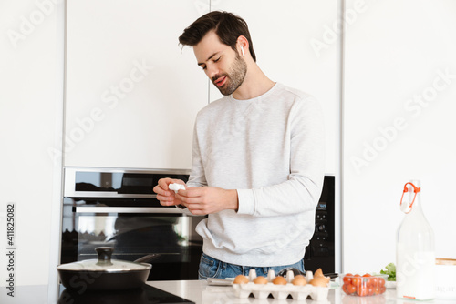 Pleased young man using earphones while cooking scrambled eggs