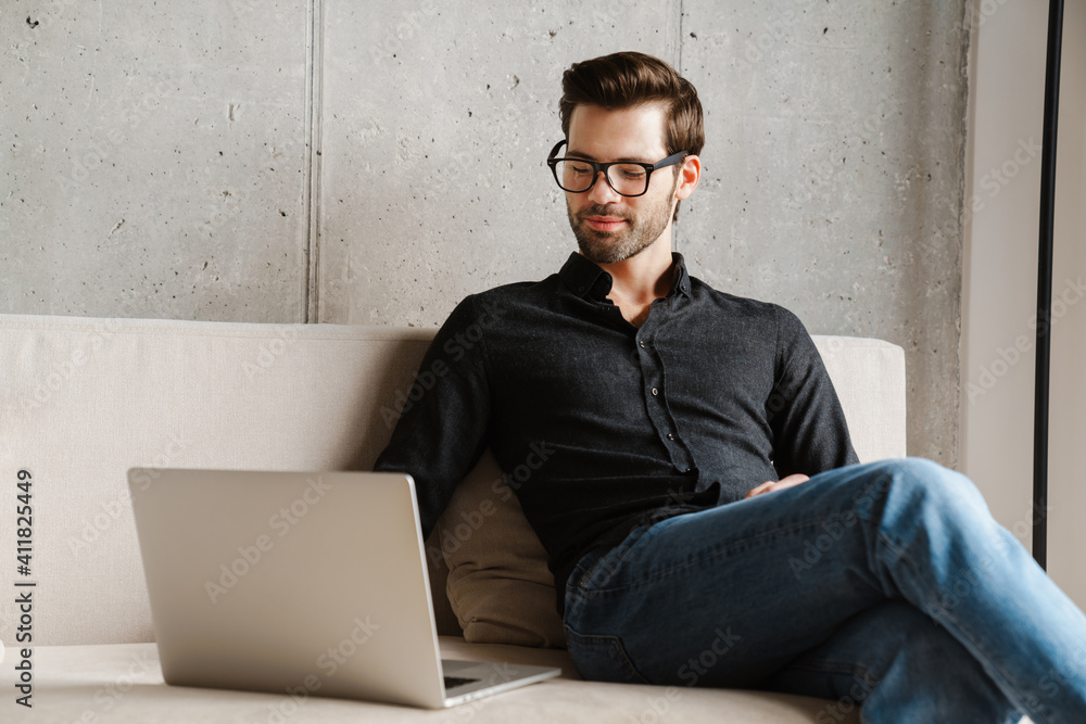 Pleased unshaven man using laptop while sitting on couch at home Pleased unshaven man using laptop while sitting on couch at home