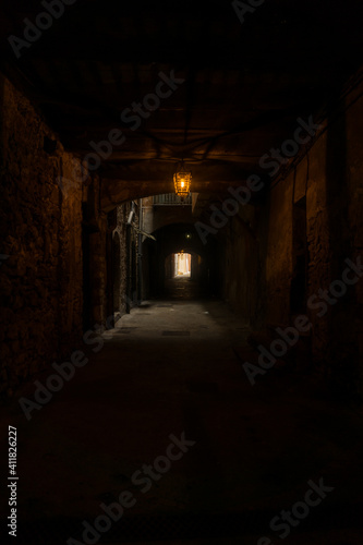Narrow and dark alley with a lantern illuminating the middle area and a door that gives light to the background. 