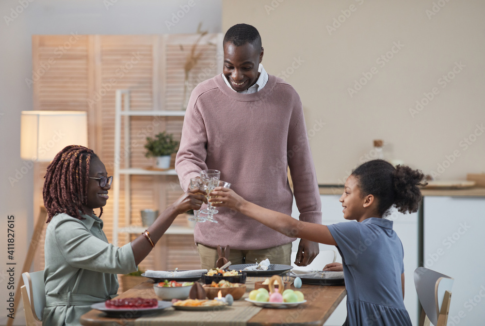 Portrait of modern African -American family clinking glasses and enjoying dinner together while celebrating Easter at home, copy space