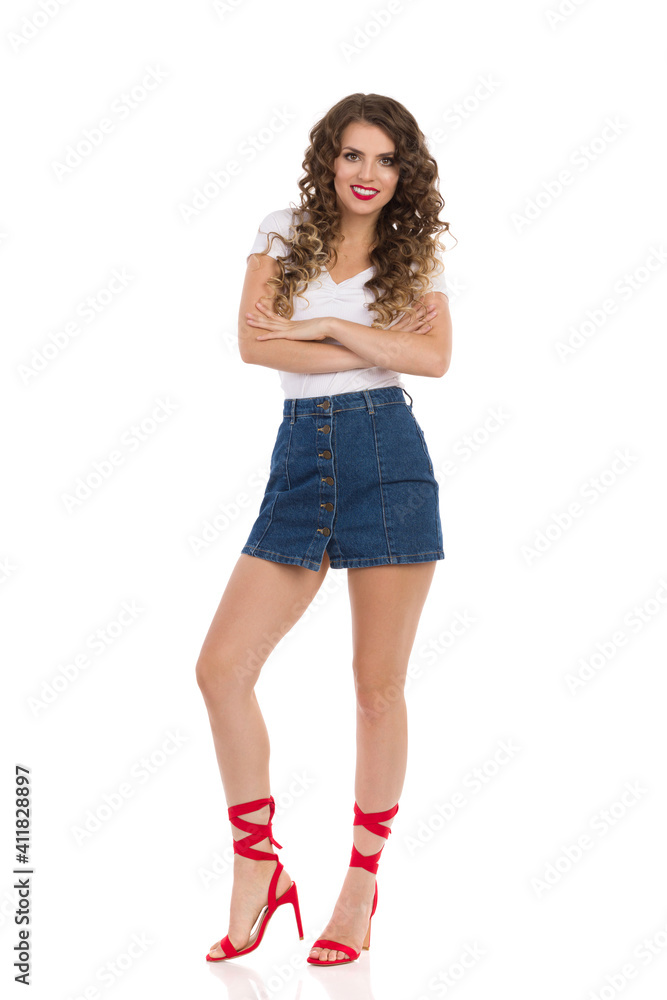 Smiling Young Woman In Jeans Mini Skirt And Red High Heels Is Standing With Arms Crossed. Smiling Young Woman In Jeans Mini Skirt And Red High Heels Is Standing With Arms Crossed.