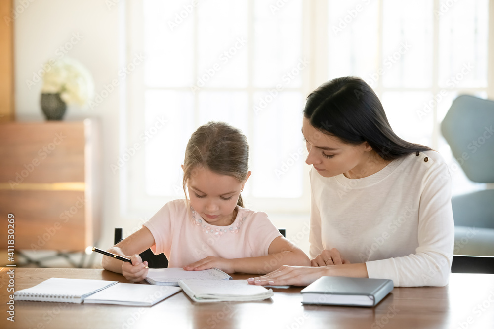 Caring young Caucasian mom and small 7s daughter sit at table at home handwriting make homework assignment together. Loving mother or nanny and little girl child study learn do home task. Caring young Caucasian mom and small 7s daughter sit at table at home handwriting make homework assignment together. Loving mother or nanny and little girl child study learn do home task.