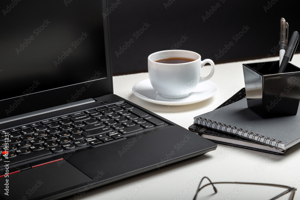 Black laptop on white table black background. Glasses, cup of coffee, office supplies. Home workspace for work or study in Office interior Black laptop on white table black background. Glasses, cup of coffee, office supplies. Home workspace for work or study in Office interior